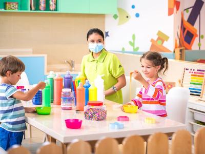Dos niños y una cuidadora hacen manualidades en una mesa dentro de un aula colorida.