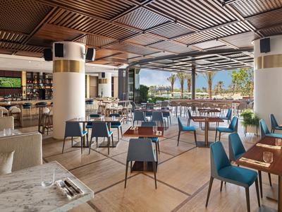 Moderne offene Hotelbar mit Holzdecke, blauen Stühlen und Blick auf Palmen und Meer.