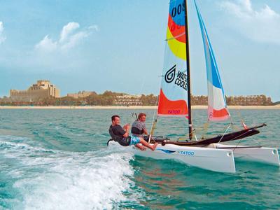 Zwei Personen segeln auf einem kleinen Katamaran bei klarem Himmel und ruhigem Meer.