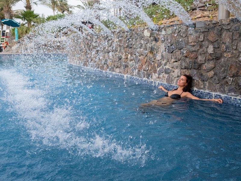 Mujer relajándose en la piscina del hotel con pared de piedra y fuentes de agua al fondo.