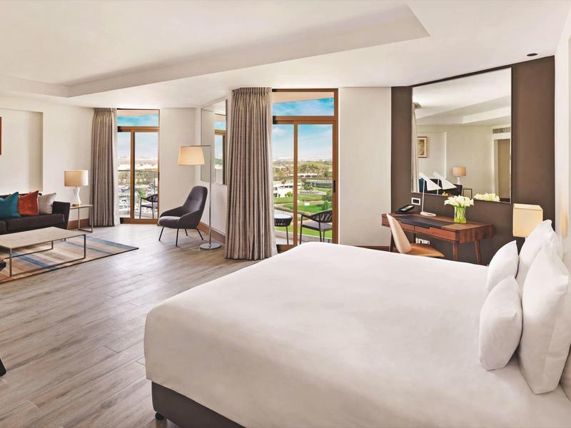 Habitación de hotel espaciosa con cama, área de estar, escritorio y balcón con vista a zonas verdes.