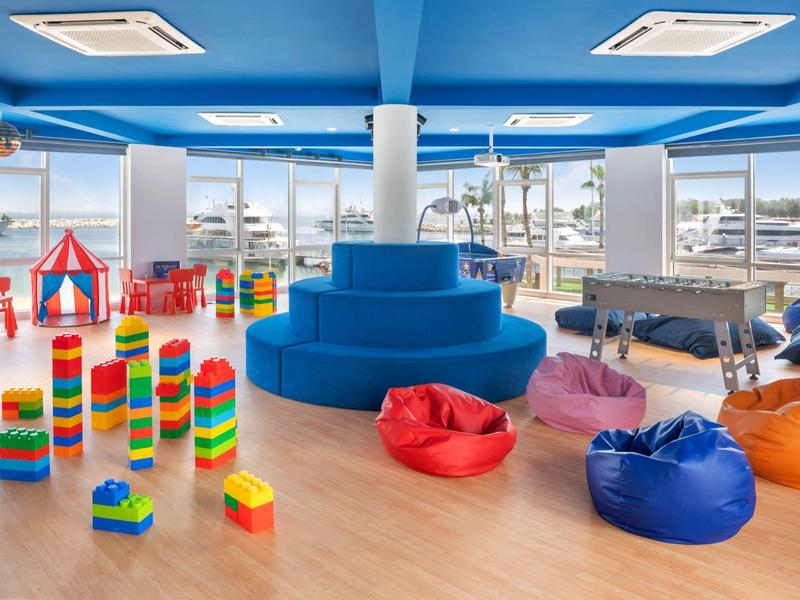 Área de juegos infantil luminosa con bloques de colores, puffs y gran asiento azul junto a ventanas.