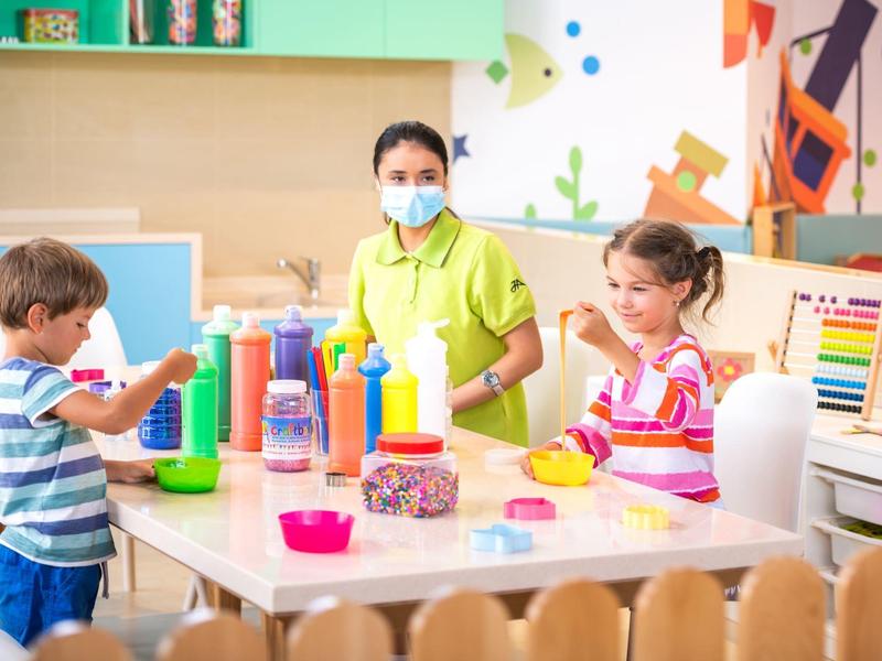 Dos niños y una cuidadora hacen manualidades en una mesa dentro de un aula colorida.