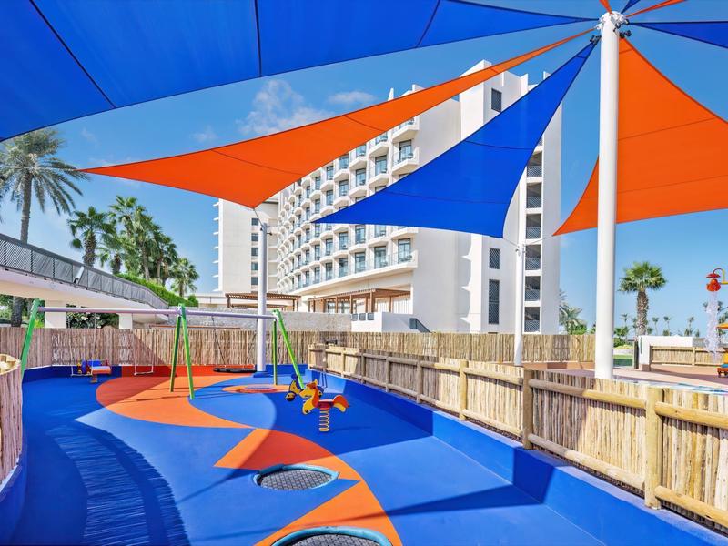 Zona de juegos al aire libre con suelo acolchonado azul y naranja y toldos de colores en un hotel.