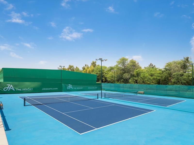 Una pista de tenis azul al aire libre con vallados verdes y árboles al fondo bajo un cielo azul.
