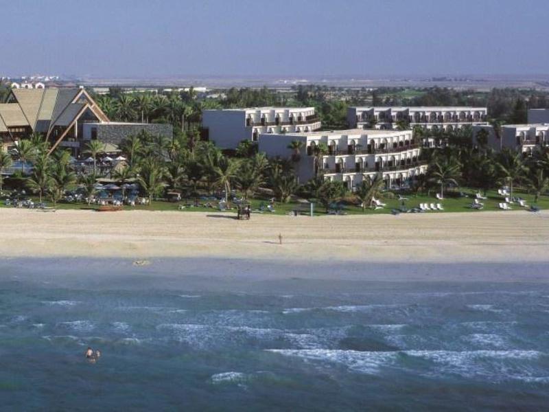 Playa de arena clara con complejo hotelero tropical con varios edificios y palmeras detrás.