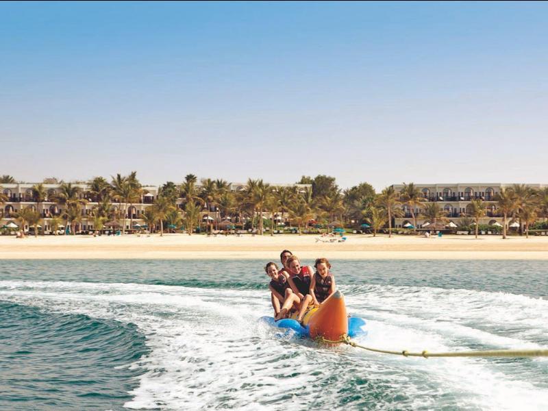 Dos personas montan un bote inflable en forma de plátano sobre el mar cerca de una playa de arena.