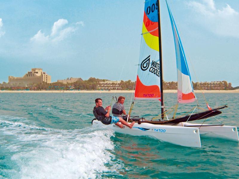 Velero con vela colorida en agua azul frente a paisaje costero bajo cielo despejado.