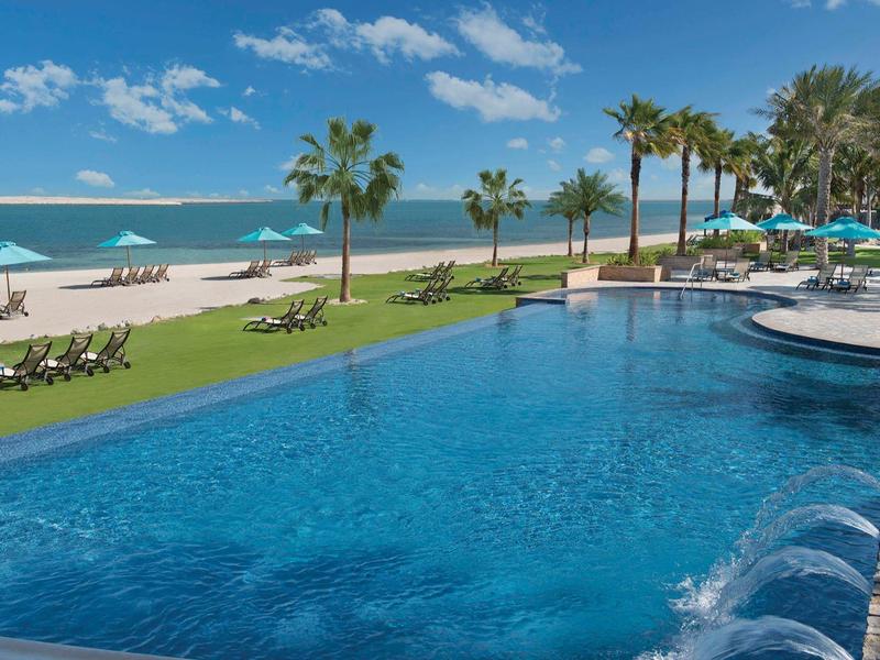 Gran piscina junto a palmeras y playa con cielo azul y mar al fondo.