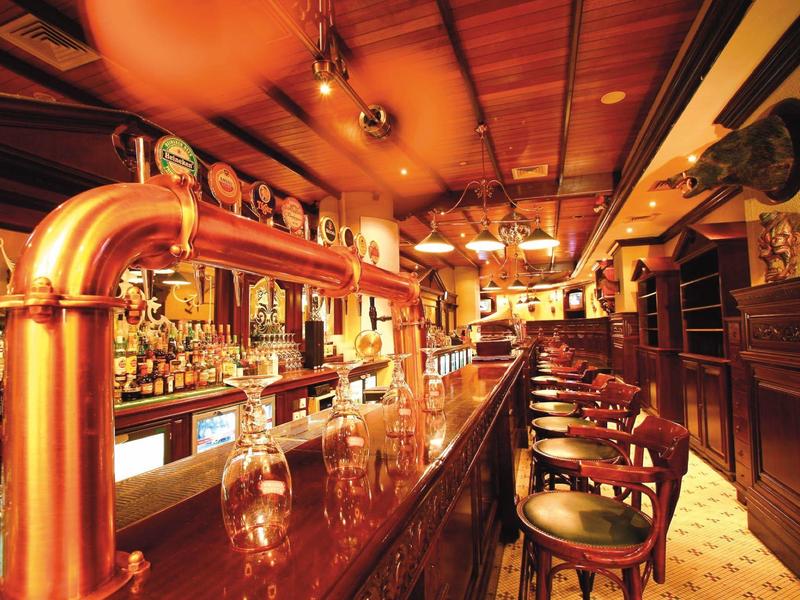 Innenansicht einer Bar mit langem Holztresen, hohen Stühlen und warmem, gelblich-rotem Licht.