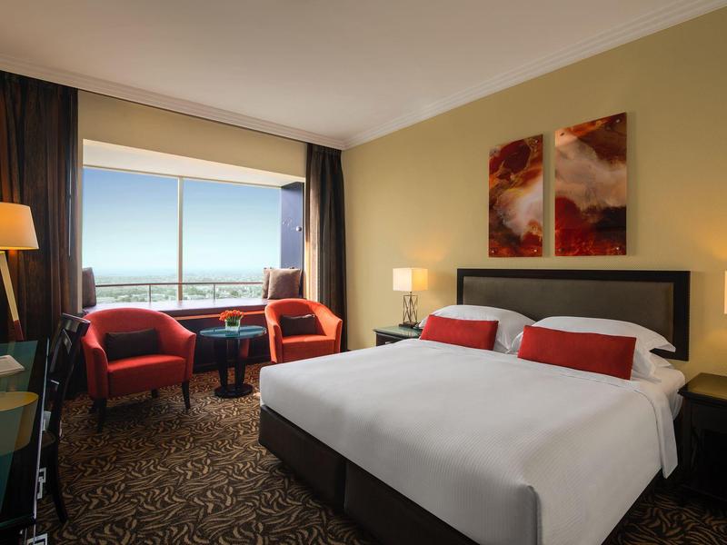 Modernes Hotelzimmer mit großem Bett, roten Sesseln, Teppichboden und Blick durch Fenster.