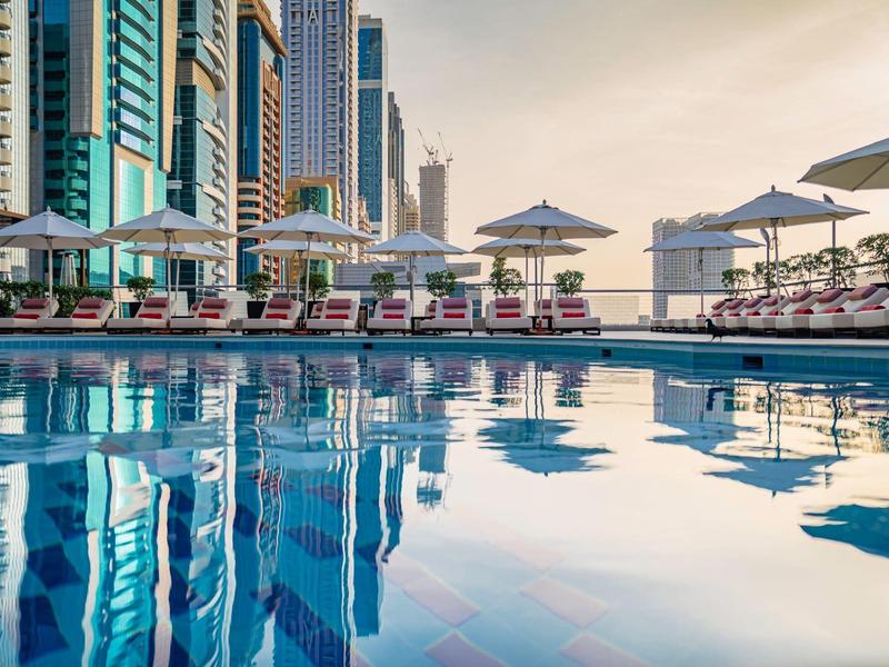 Blauer Pool mit roten Liegestühlen und weißen Sonnenschirmen vor hohen Glasgebäuden.