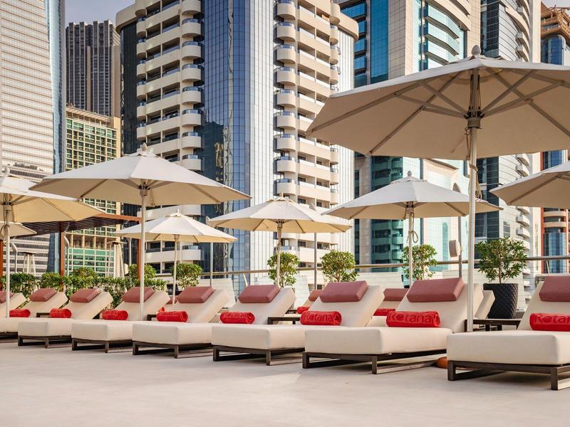 Reihe bequemer Liegestühle mit roten Kissen unter großen Sonnenschirmen neben Hochhäusern.
