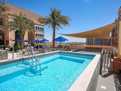 Piscina moderna de hotel con sombrillas azules y palmeras bajo un cielo despejado.