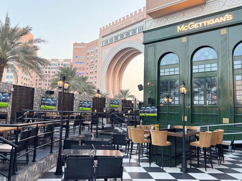 Área exterior de un café con mesas y sillas, palmeras y un gran arco al fondo.