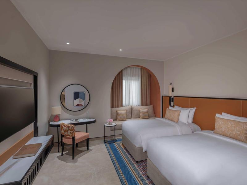 Habitación de hotel moderna con dos camas individuales, zona de estar y ventana grande con cortinas.