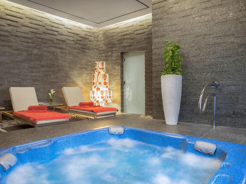 Jacuzzi iluminado con burbujas en sala con paredes de piedra y tumbonas con toallas rojas.