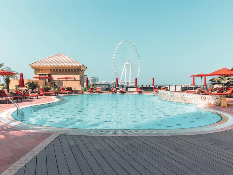 Piscina redonda al aire libre en hotel con sombrillas rojas y sillas bajo cielo claro.