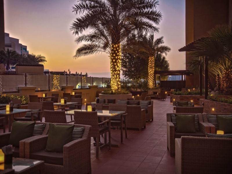 Terraza del hotel al atardecer con sillas, mesas y palmeras iluminadas al fondo.