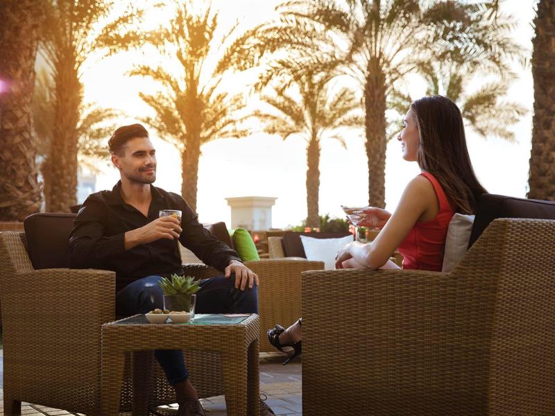 Dos personas sentadas en sillones de mimbre conversan al aire libre al atardecer junto a palmeras.