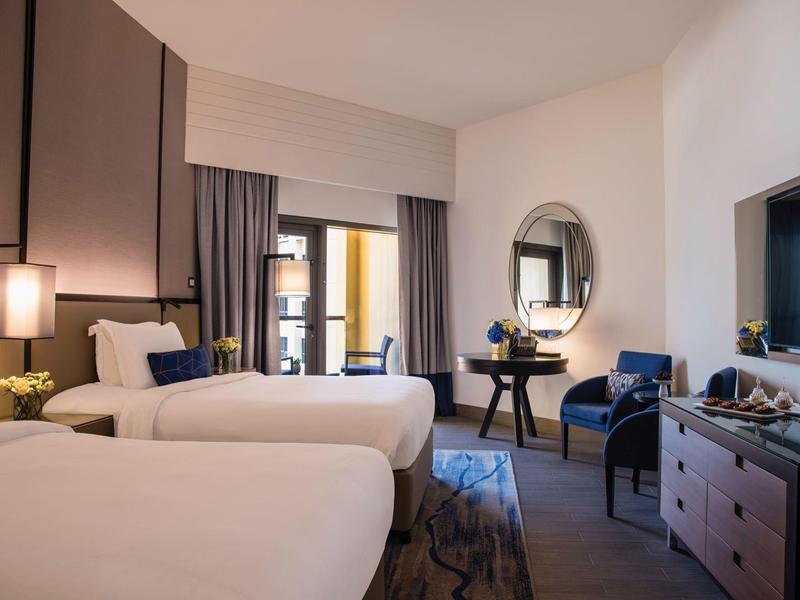 Habitación de hotel moderna con dos camas, mesa con silla y televisión, decorada en tonos neutros.