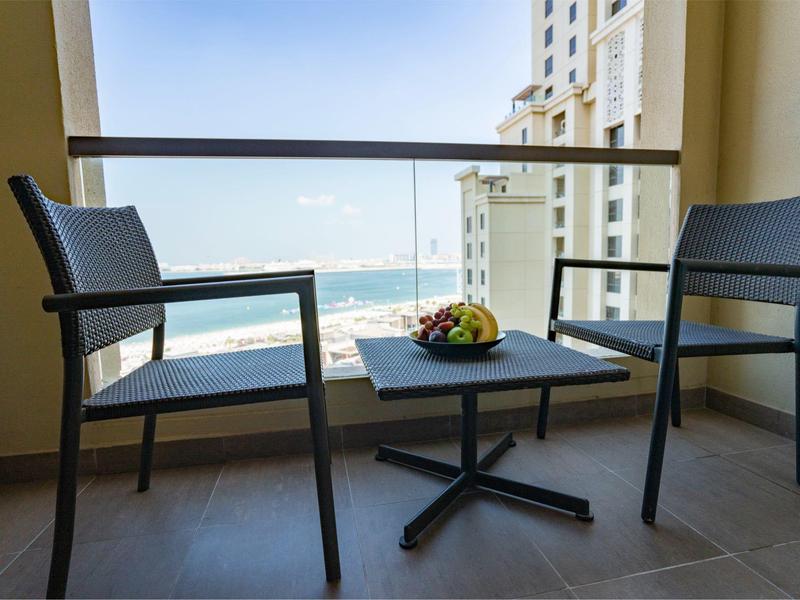 Balcón con dos sillas y una mesa con una bandeja de fruta, vista al mar y edificios.