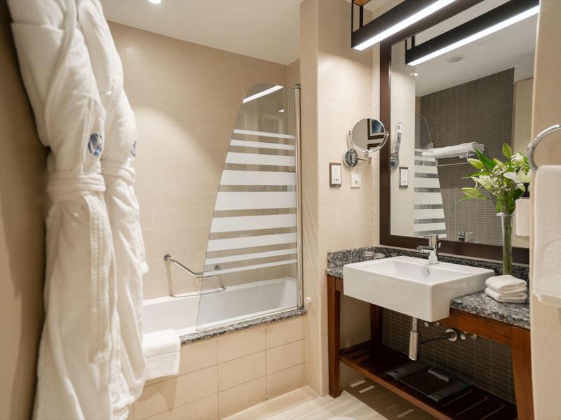 Baño moderno con bañera, toallas, albornoces y lavabo con espejo iluminado.