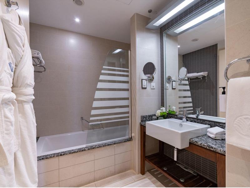 Baño de hotel moderno con ducha, bañera, espejo grande y albornoces blancos.