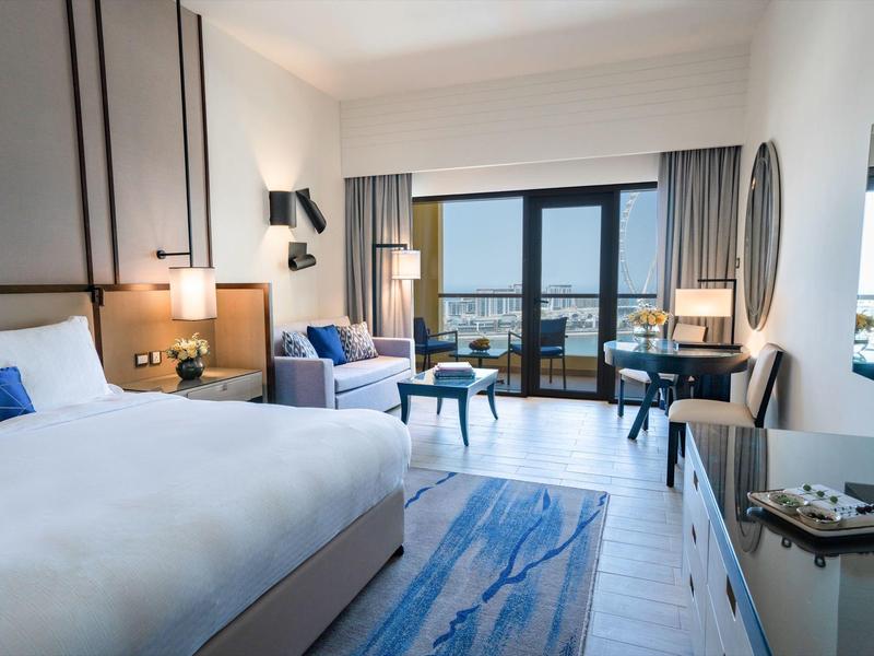 Habitación de hotel moderna con cama doble, sofá, escritorio y balcón con vista al mar.