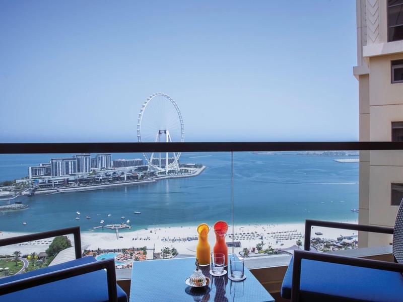 Vista desde un balcón con barandilla de vidrio hacia el mar, la playa y una noria bajo un cielo despejado.