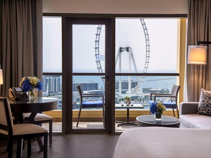 Habitación de hotel con balcón y vista a una noria junto al agua bajo cielo nublado.