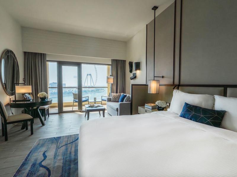 Habitación de hotel moderna con cama grande, área de estar y vista a la terraza y el agua.