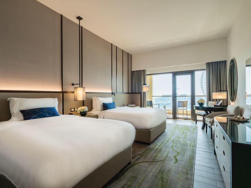 Habitación de hotel moderna con dos camas grandes, ventanal y escritorio en luz natural.