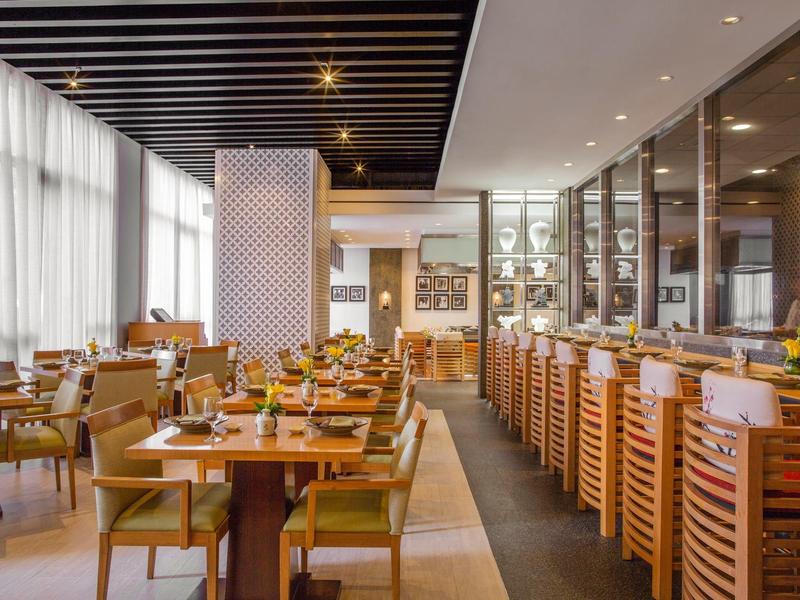 Área de restaurante de hotel moderno con mesas, sillas y grandes ventanas con mucha luz natural.