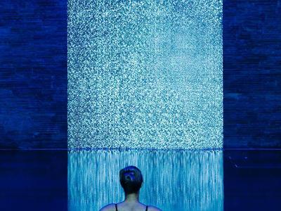 Person schwimmt in dunklem Becken vor hellem Wasserfall-Wand im blauen Licht.
