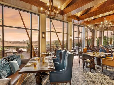 Restaurant mit großen Fenstern, blauen Sesseln, Holzbalken und Außenblick bei Sonnenuntergang.