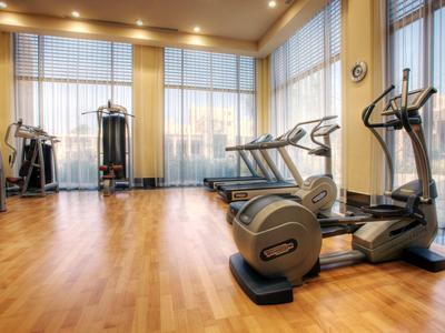 Heller Fitnessraum mit Holzfußboden, Laufbändern und Trainingsgeräten vor großen Fenstern.