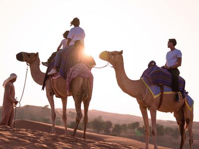 Zwei Personen reiten auf Kamelen in einer Wüstenlandschaft bei Sonnenuntergang.