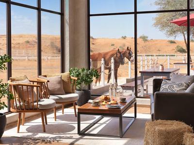 Helle Hotellounge mit großen Fenstern zeigt Wüstenlandschaft und Kamele draußen.