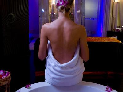 Frau mit nassem Haar, in weißem Handtuch, vor Badewanne mit Rosenblättern, gedimmtes blaues Licht.