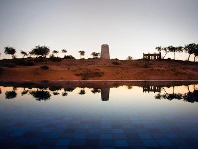 Schatten von Bäumen und Turm spiegeln sich ruhig in klarem, blauem Wasser bei Sonnenuntergang.