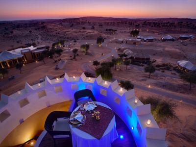 Wüstenlandschaft mit Zeltunterkünften und gedecktem Tisch auf Terrasse bei Sonnenuntergang.