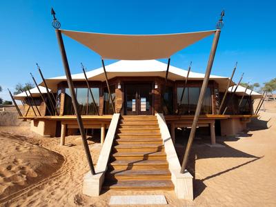 Modernes Gebäude mit Holztreppe, großem Sonnensegel und sandiger Wüstenumgebung bei klarem Himmel.