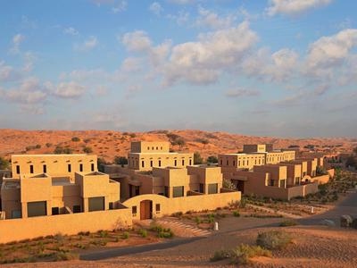 Wohnhäuser in heller Sandsteinfarbe liegen in einer trockenen Wüstenlandschaft unter blauem Himmel.