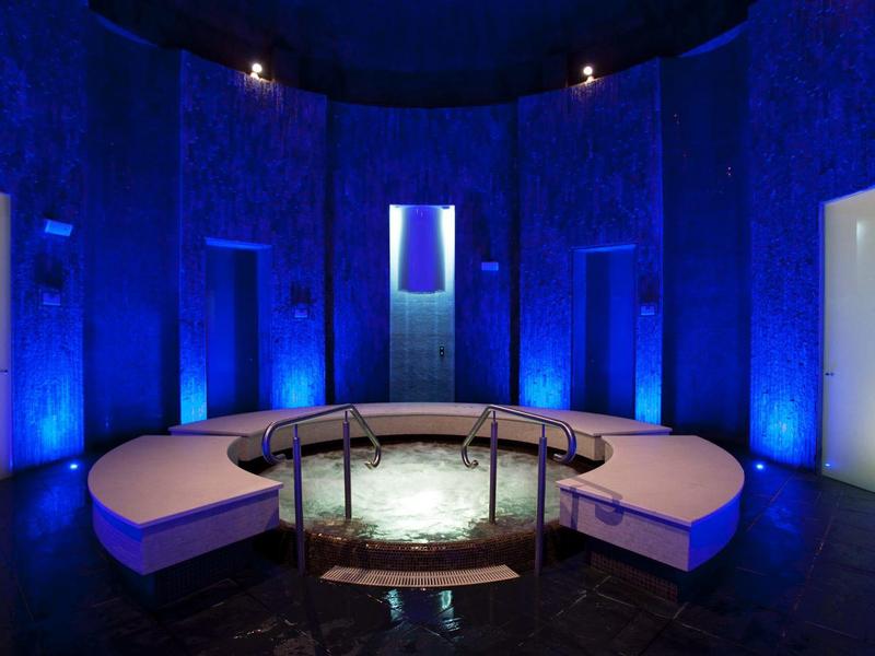 Runder Raum mit blauen Wänden, beleuchteter Wasserfläche und weißen Sitzbänken mit Geländer.