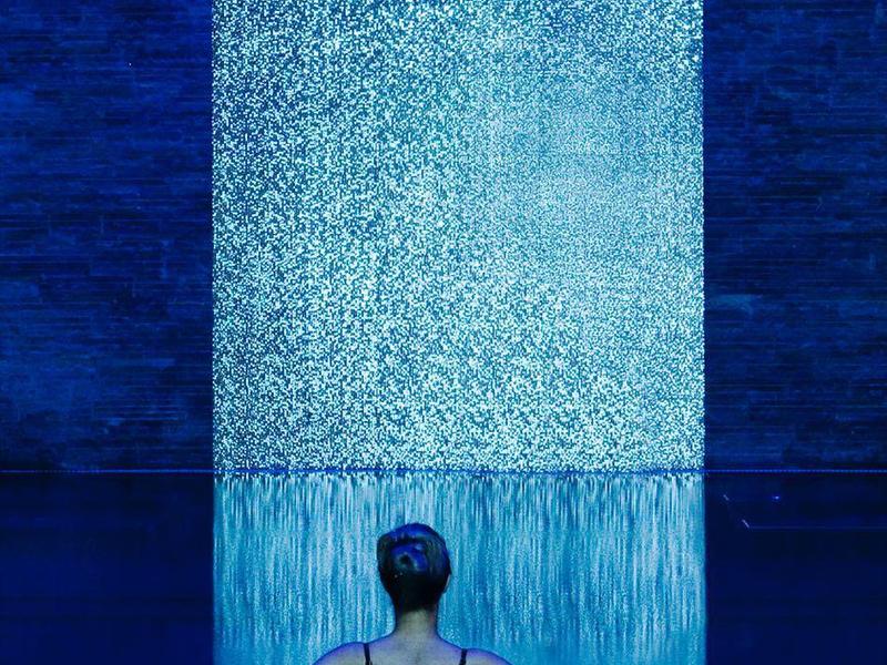 Person schwimmt in dunklem Becken vor hellem Wasserfall-Wand im blauen Licht.