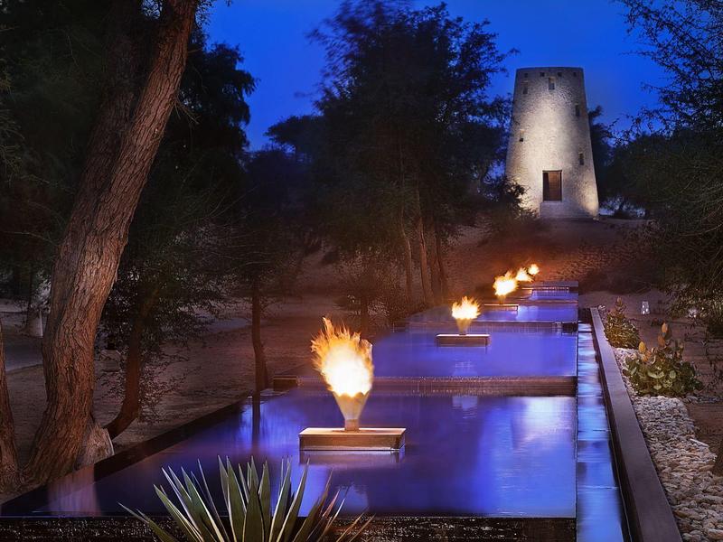 Beleuchteter Wasserbrunnen mit Flammen und Pflanzen bei Nacht vor einem Turm.
