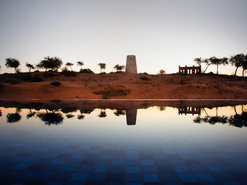Schatten von Bäumen und Turm spiegeln sich ruhig in klarem, blauem Wasser bei Sonnenuntergang.