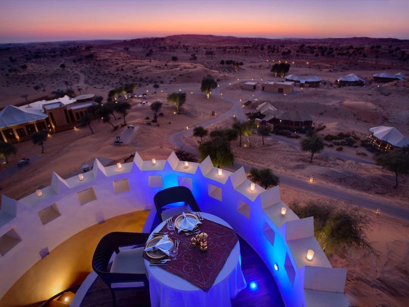 Wüstenlandschaft mit Zeltunterkünften und gedecktem Tisch auf Terrasse bei Sonnenuntergang.