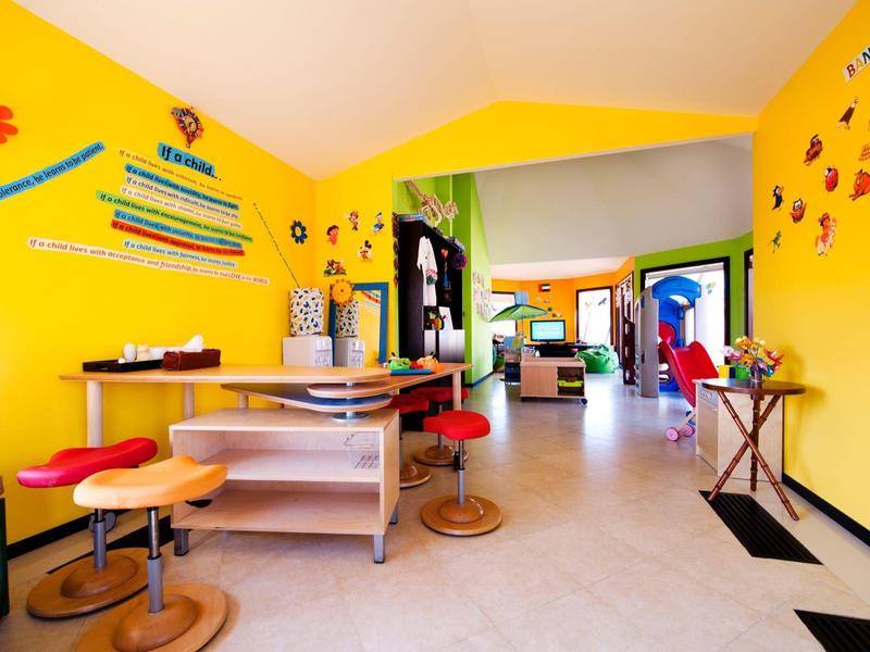 Bunter Kindergartenraum mit gelben Wänden, niedrigen Tischen, bunten Hockern und Kinderdeko.